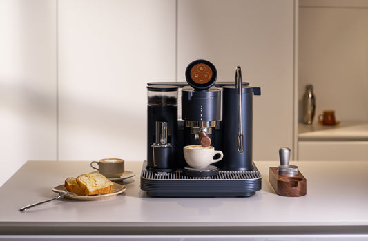The image shows a high-end espresso machine with various accessories like a tamper, grinder, and cups, all arranged on a counter. The machine has a sleek, modern design and appears to be part of a well-equipped home or cafe setup.