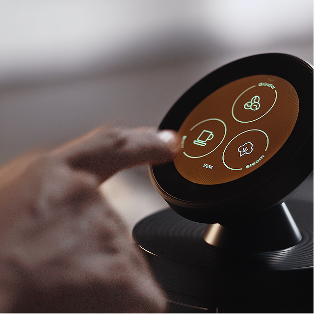 Close-up of a hand using the Meraki espresso machine’s circular touchscreen, showing grinder, brewing and steam controls.