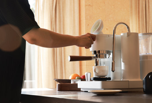Person using sleek white espresso machine with wooden handle to brew coffee into white cup on kitchen counter