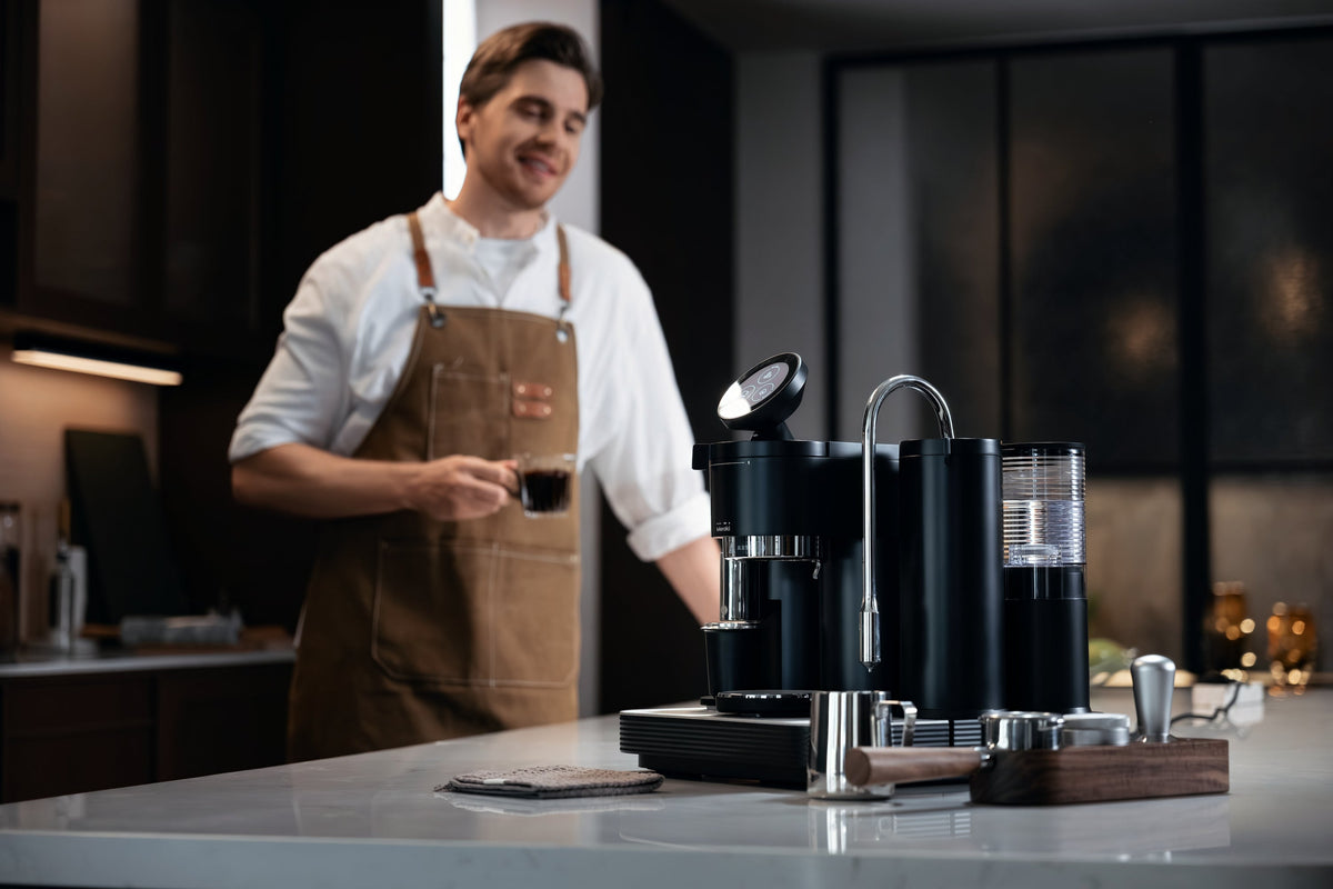 Person wearing a brown apron holding a glass of coffee beside a Meraki espresso machine and portafilter tools.