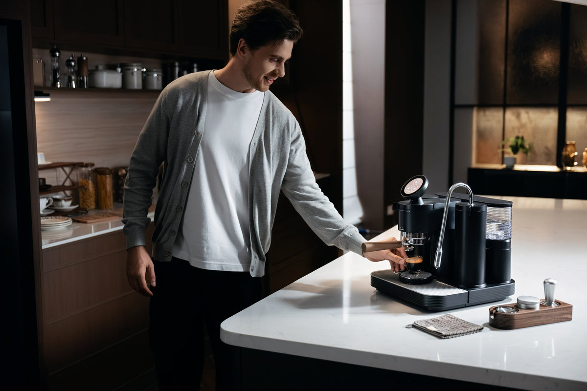 Man reaching for a glass of espresso beneath the portafilter of a black Meraki espresso machine
