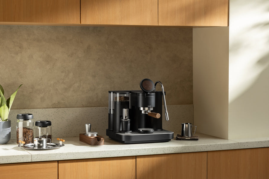 Black Meraki espresso machine with built-in grinder on a kitchen counter beside coffee beans, preparation tools and a milk pitcher.