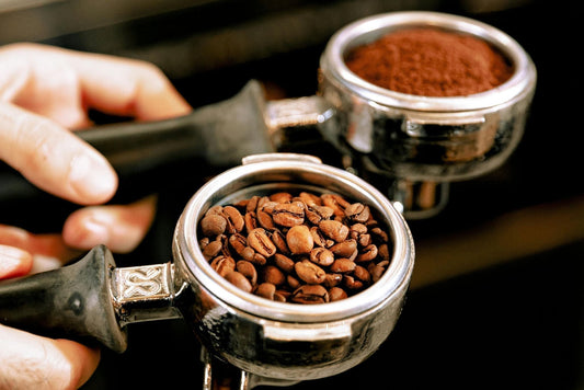 Two espresso portafilters held in hands; one filled with whole roasted coffee beans and the other with fine coffee grounds