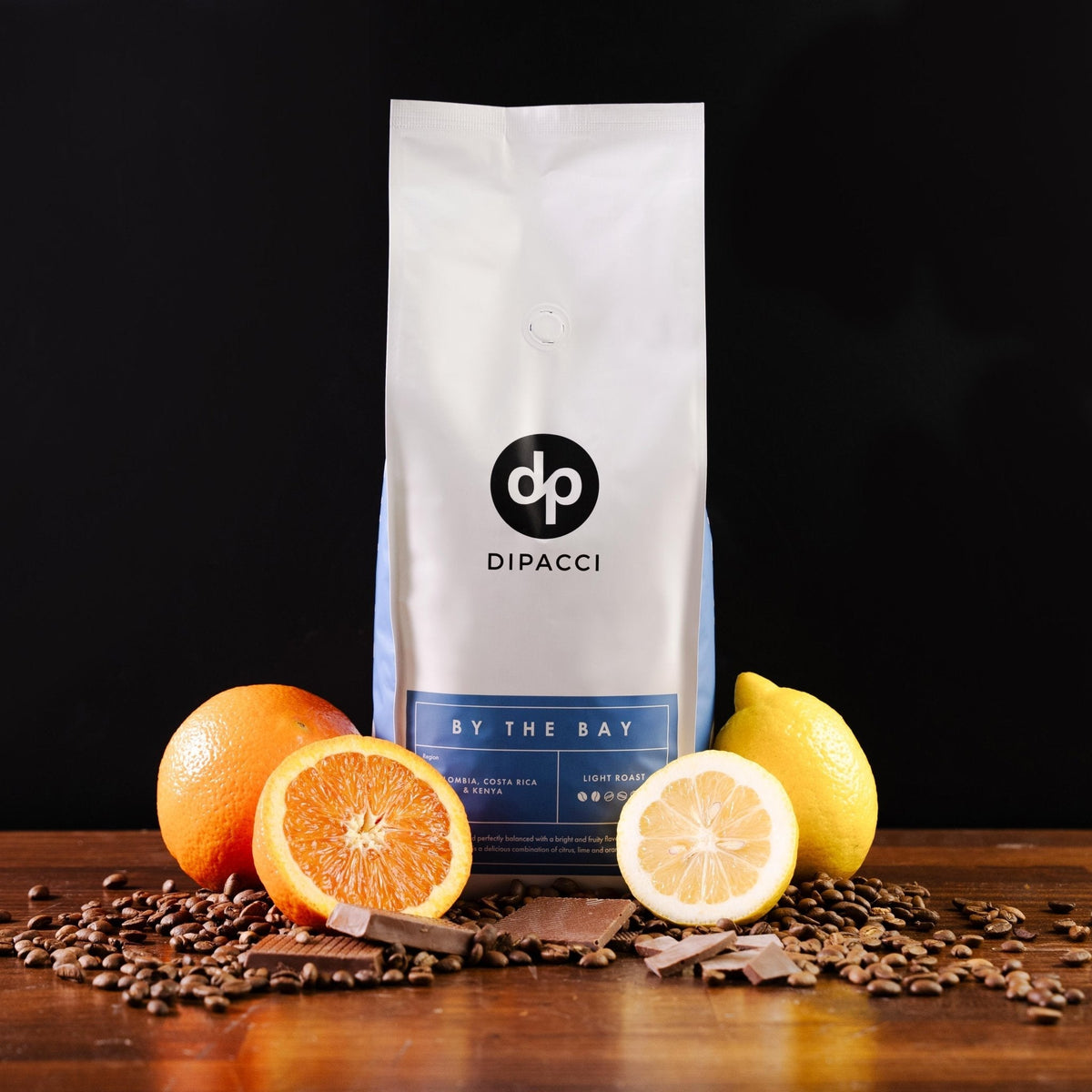 By The Bay light roast coffee bag with oranges, lemons, chocolate pieces, and coffee beans on a wooden table.