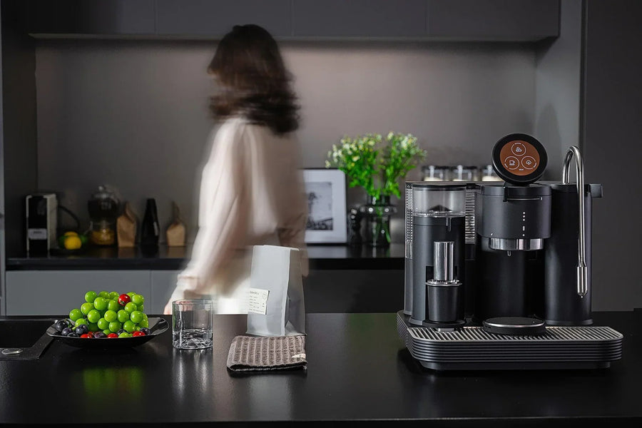 Black Meraki espresso machine with built-in grinder, circular touchscreen and steam wand on a kitchen island beside a bag of coffee beans