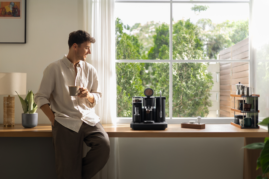 A person standing beside a black Meraki espresso machine in a sunlit kitchen