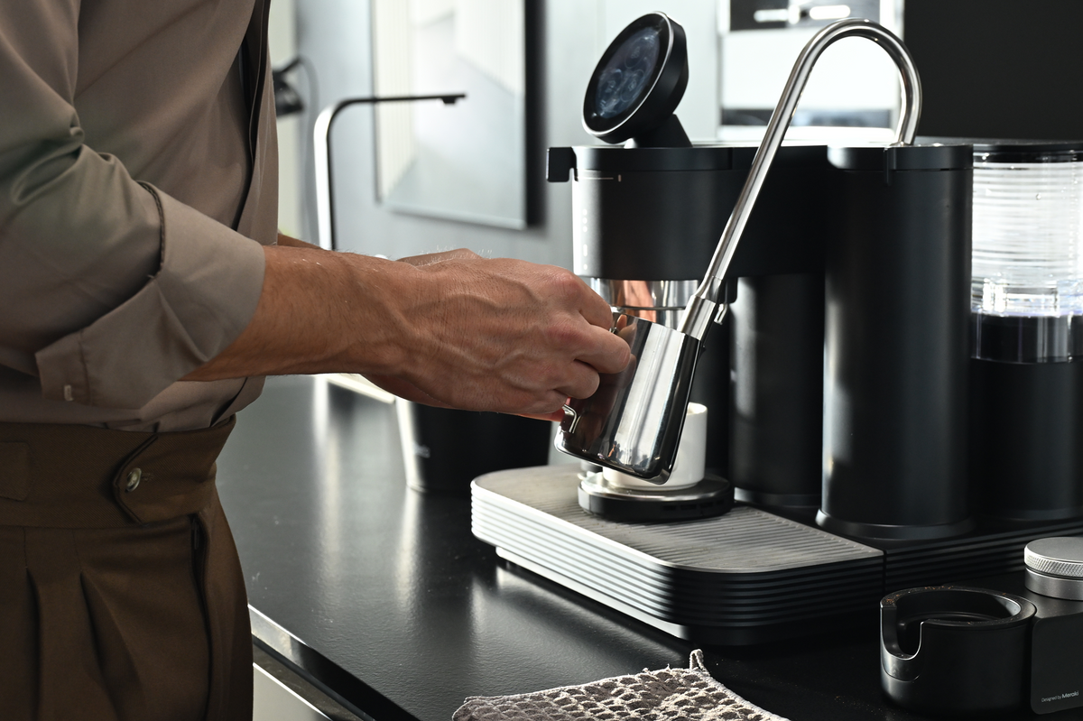 Steaming milk in a stainless steel pitcher with a black Meraki espresso machine