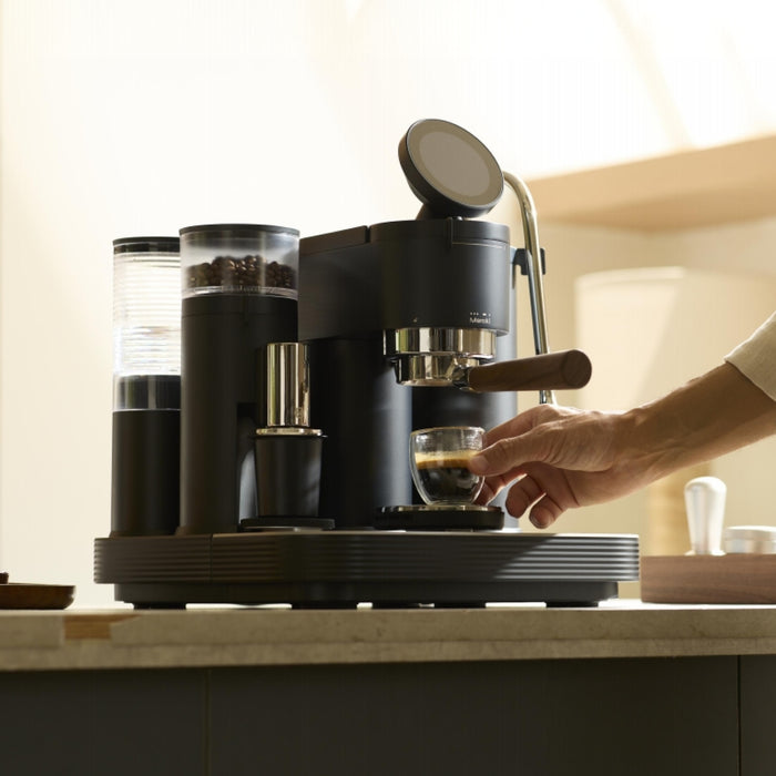 Black Meraki espresso machine with built-in grinder on a kitchen counter, hand picking up a freshly pulled espresso